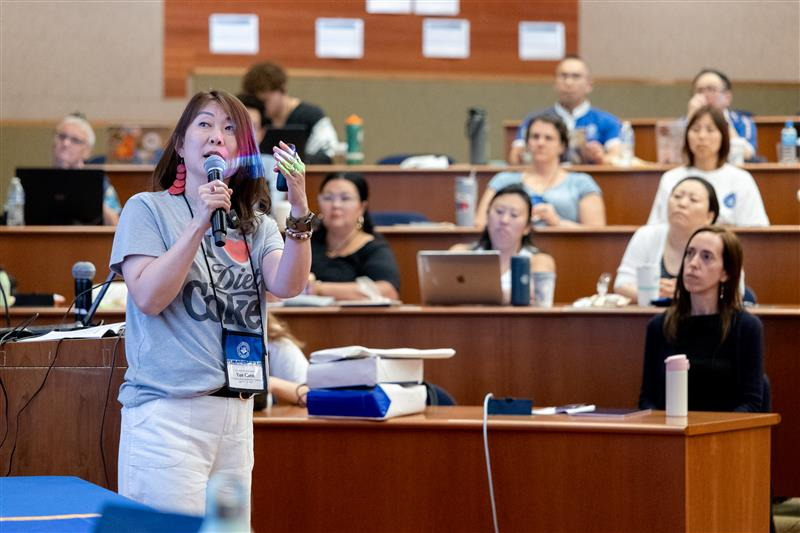 A woman speaks into the microphone and gestures at a projector screen in front of an auditorium of people.