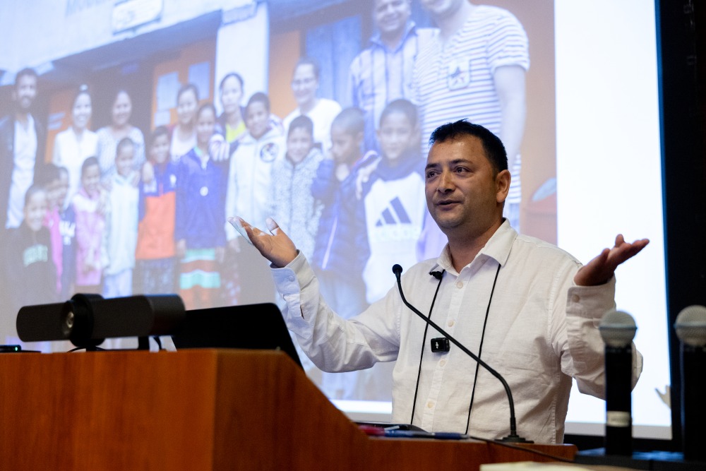 A man wearing a white button down shirt gestures with both palms up as he stands in front of a projector screen and delivers a speech.