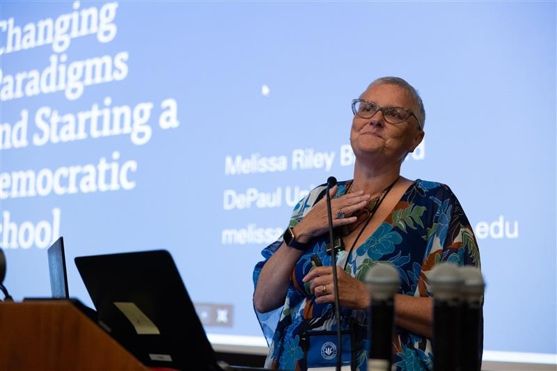 A short-haired woman, wearing a floral shirt and glasses holds her hand to her heart as she stands in front of a projector screen and delivers a keynote talk.