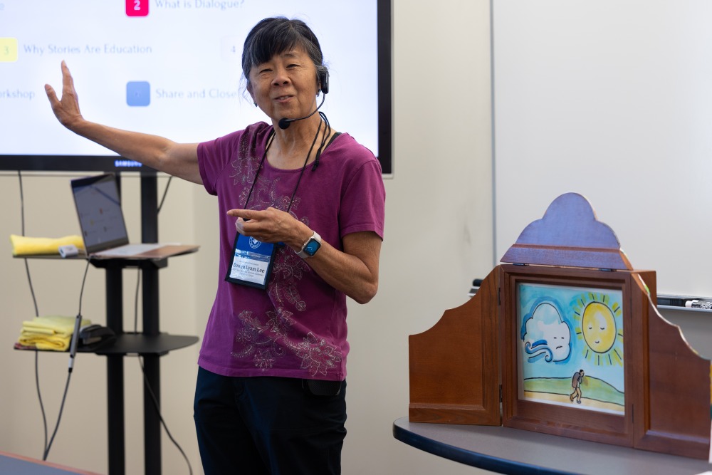 A woman wearing a microphone gestures towards the projector screen as she gives a presentation.