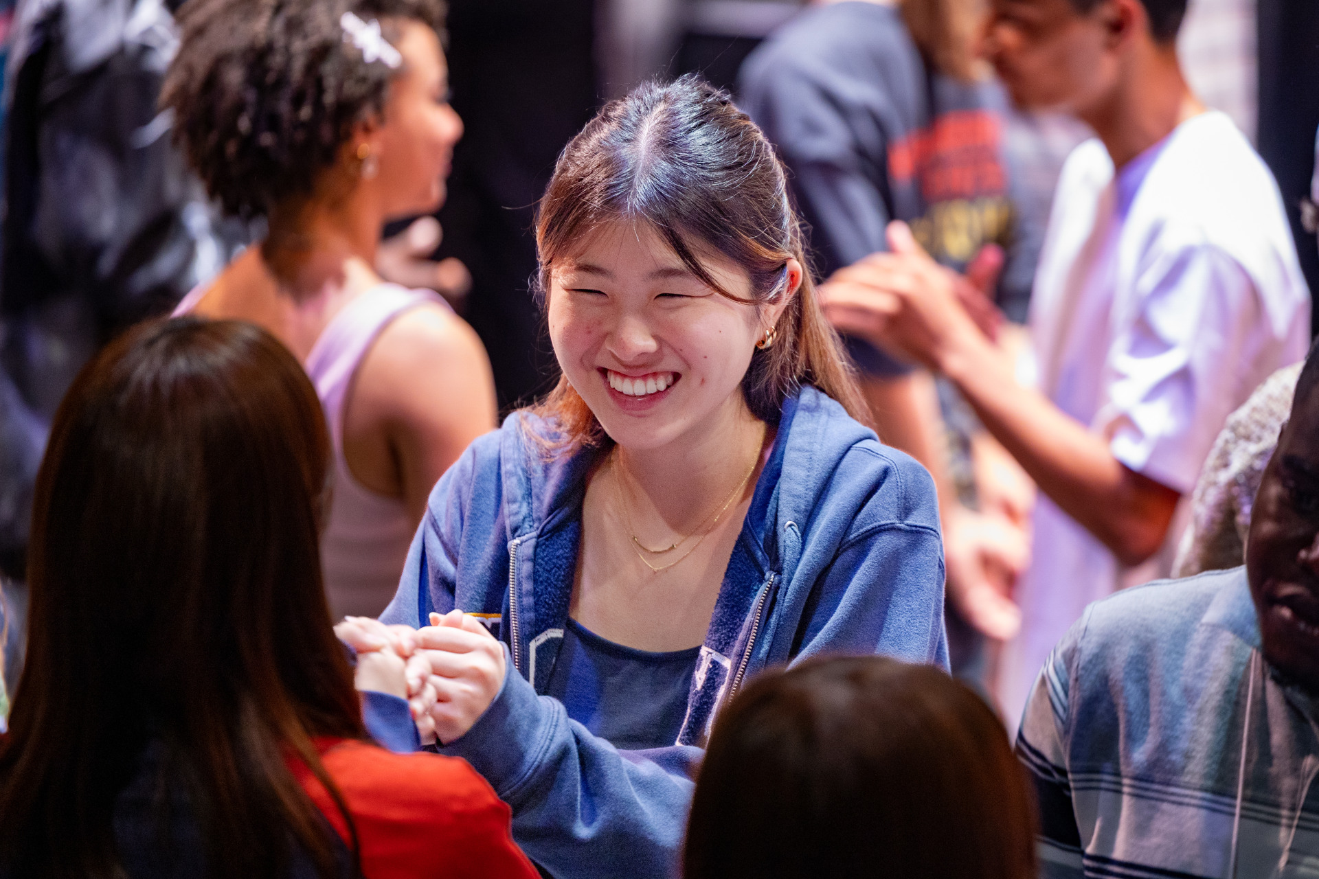 A girl wearing a blue zip-up sweater smiles at another student as they participate in an icebreaker activity.