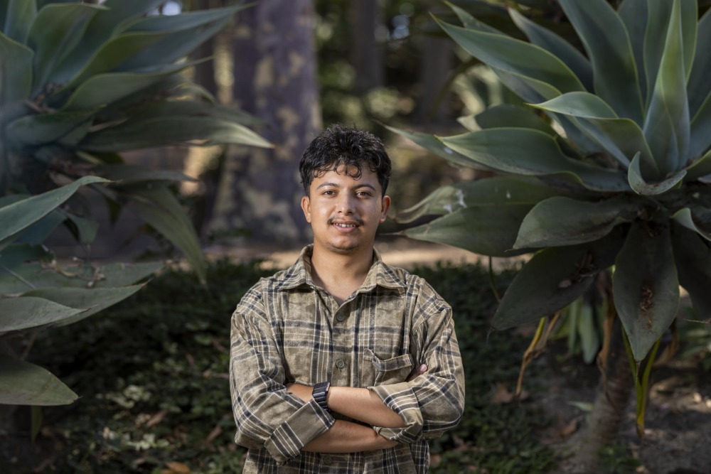 Aakash Baral, wearing a green and black plaid shirt, crosses his arms and smiles while posing by plants on SUA's campus.