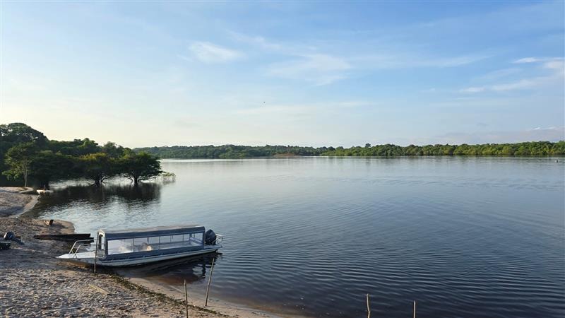 A boat is docked on the sand lining a large body of water surrounded by green trees.