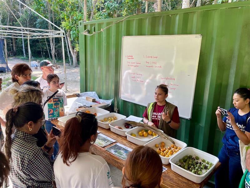Students gather around a table containing white bins full of various fruits and plants while listening to a staff member speak in front of a white board with red text.