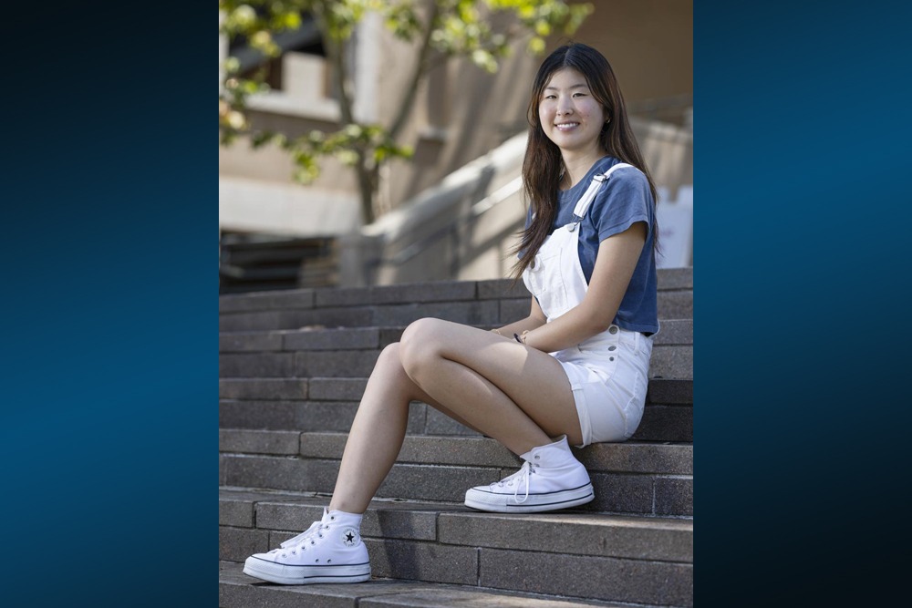 Lana Ikeda, wearing a blue t-shirt with white short overalls and white converse hightops, sits on stairs on SUA's campus.