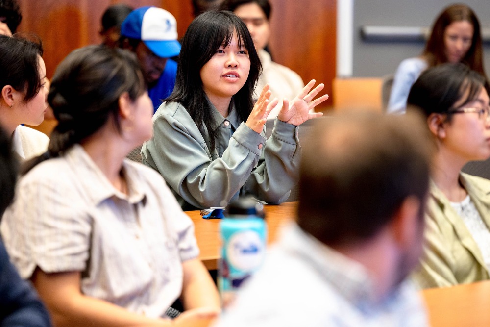 A female student gestures with her hands as she speaks while sitting in a classroom with other students.