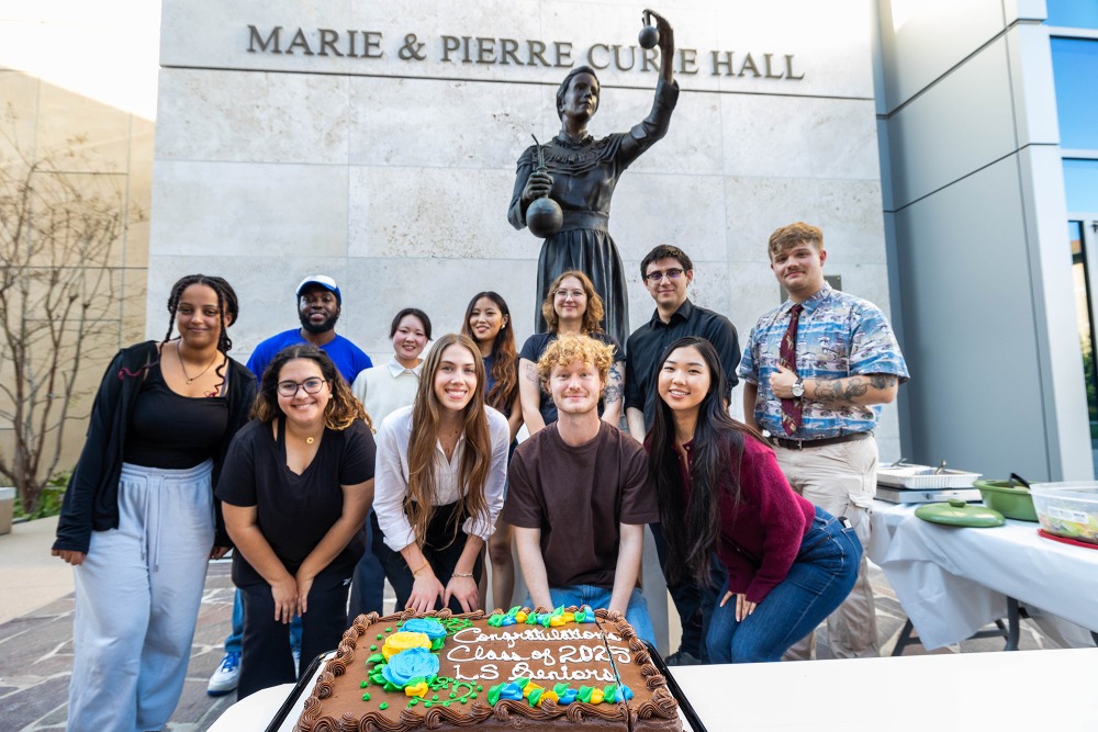 A diverse group of students smiles for a photo behind a "Congratulations LS Seniors Class of 2025" cake, standing outside Marie & Pierre Curie Hall.