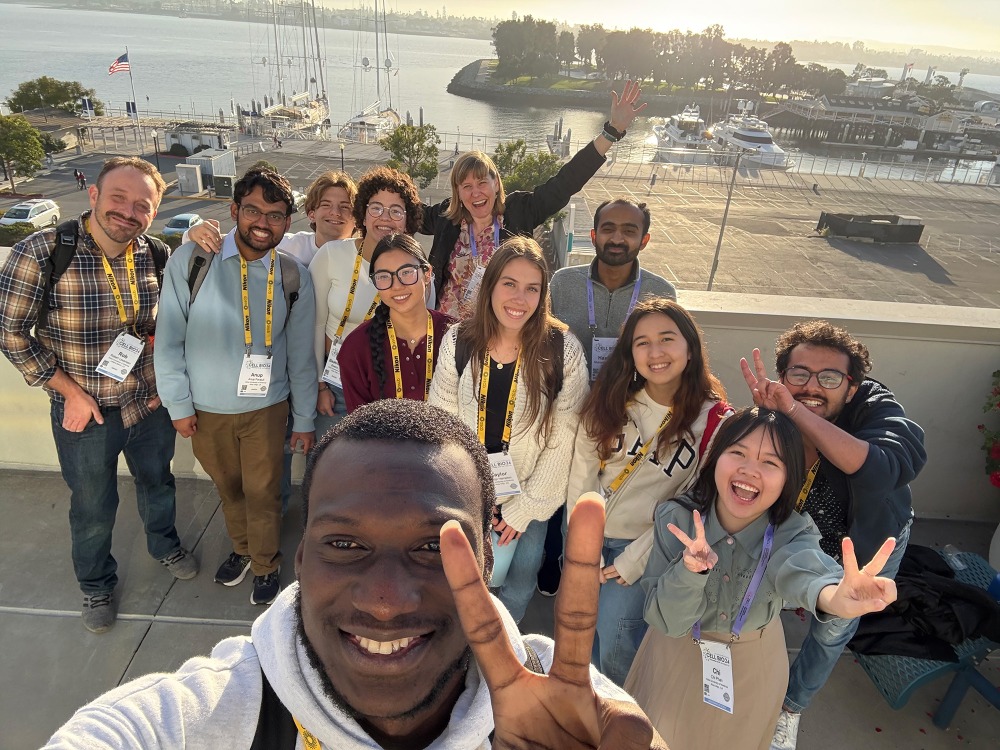 A smiling young man takes a selfie with a diverse group of people on a balcony overlooking a marina filled with boats.