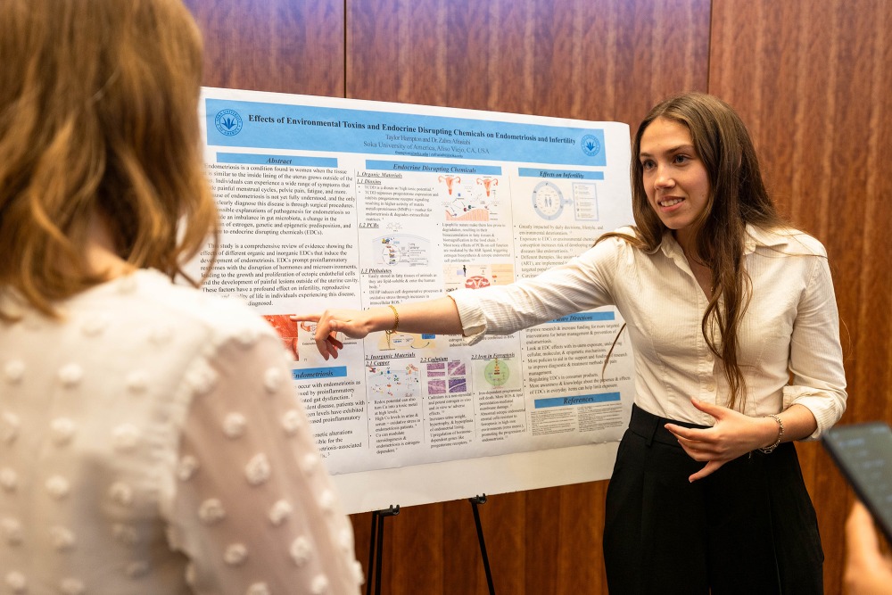 A young woman points to a scientific poster on an easel, presenting her research to another person at a Capstone presentation event.