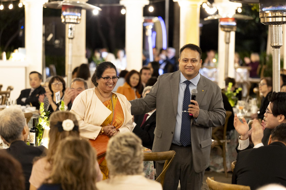 Nitesh Sil ’11, wearing a gray suit and blue tie, holds a microphone and puts his arm around a woman wearing an orange dress with a white jacket while he speaks to the gala attendees.
