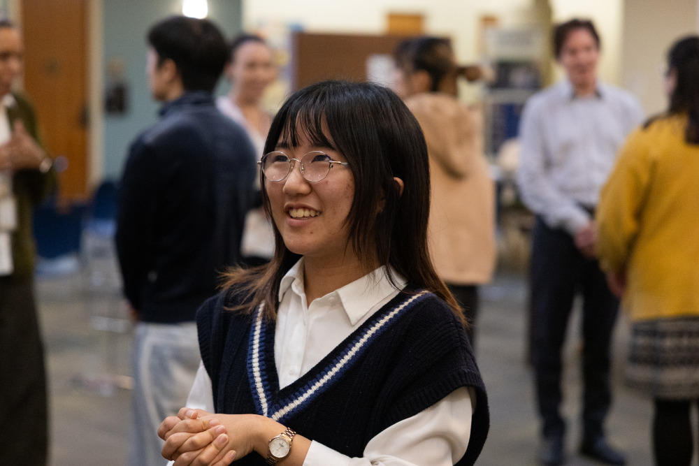A female student with black hair wearing glasses is smiling, holding her two hands together