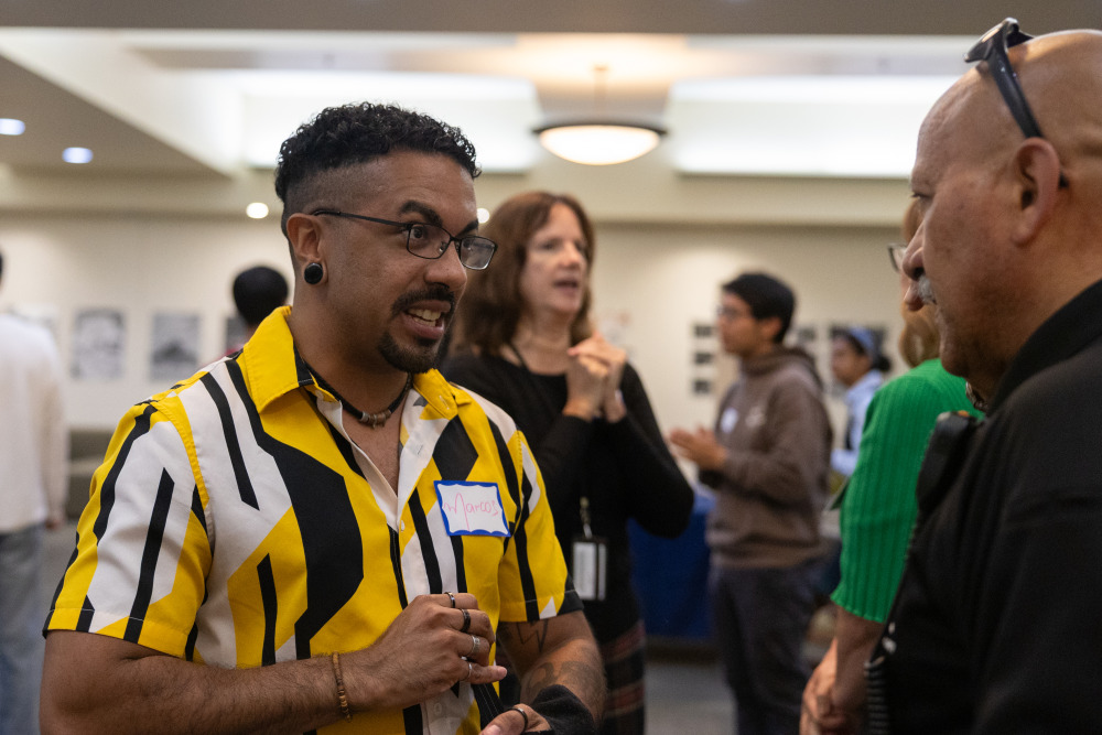 A man with short curly black hair in a yellow-black-white stripe shirt talking to another man with sunglasses