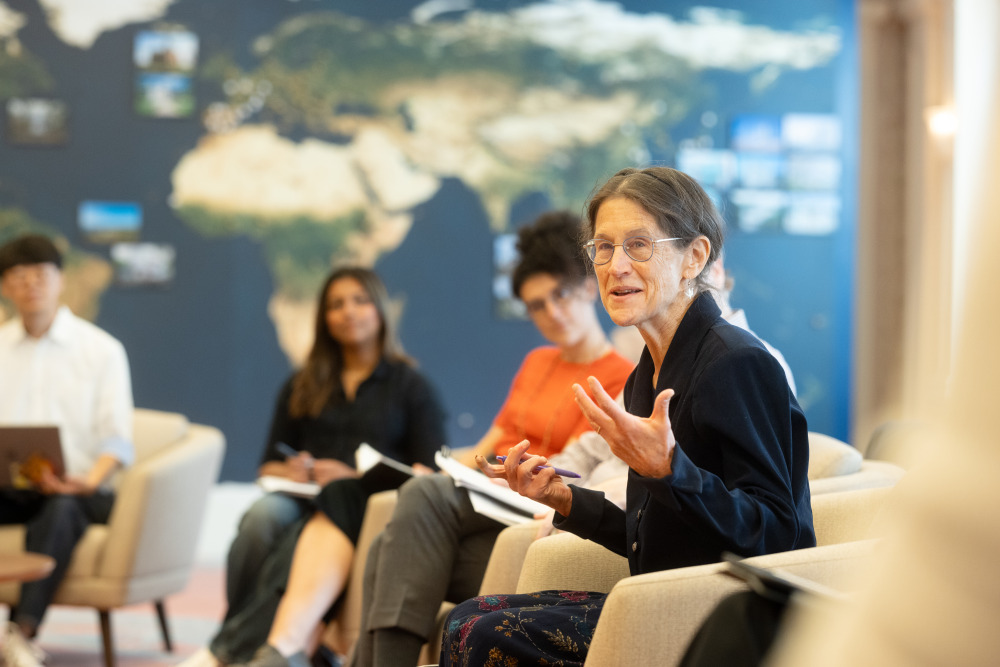 A woman with brown hair in a ponytail and wearing glasses speaks with her hands. Out-of-focus students are in the background listening sitting in front of a wall-sized navy, gold, and green world map.