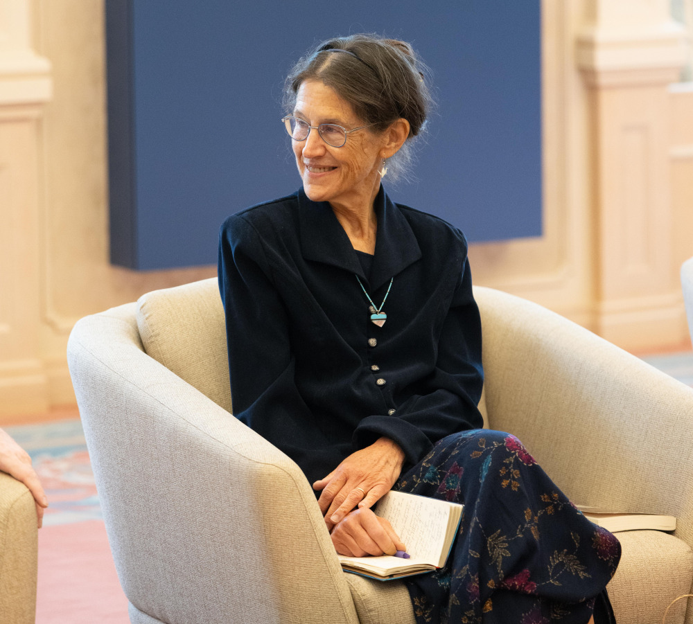 A glasses-wearing woman with her brunette hair pulled back into a bun and wearing a navy blue collared shirt and blue floral long skirt crosses her hands over an open journal and smiles to her right as she listens to someone out of frame.