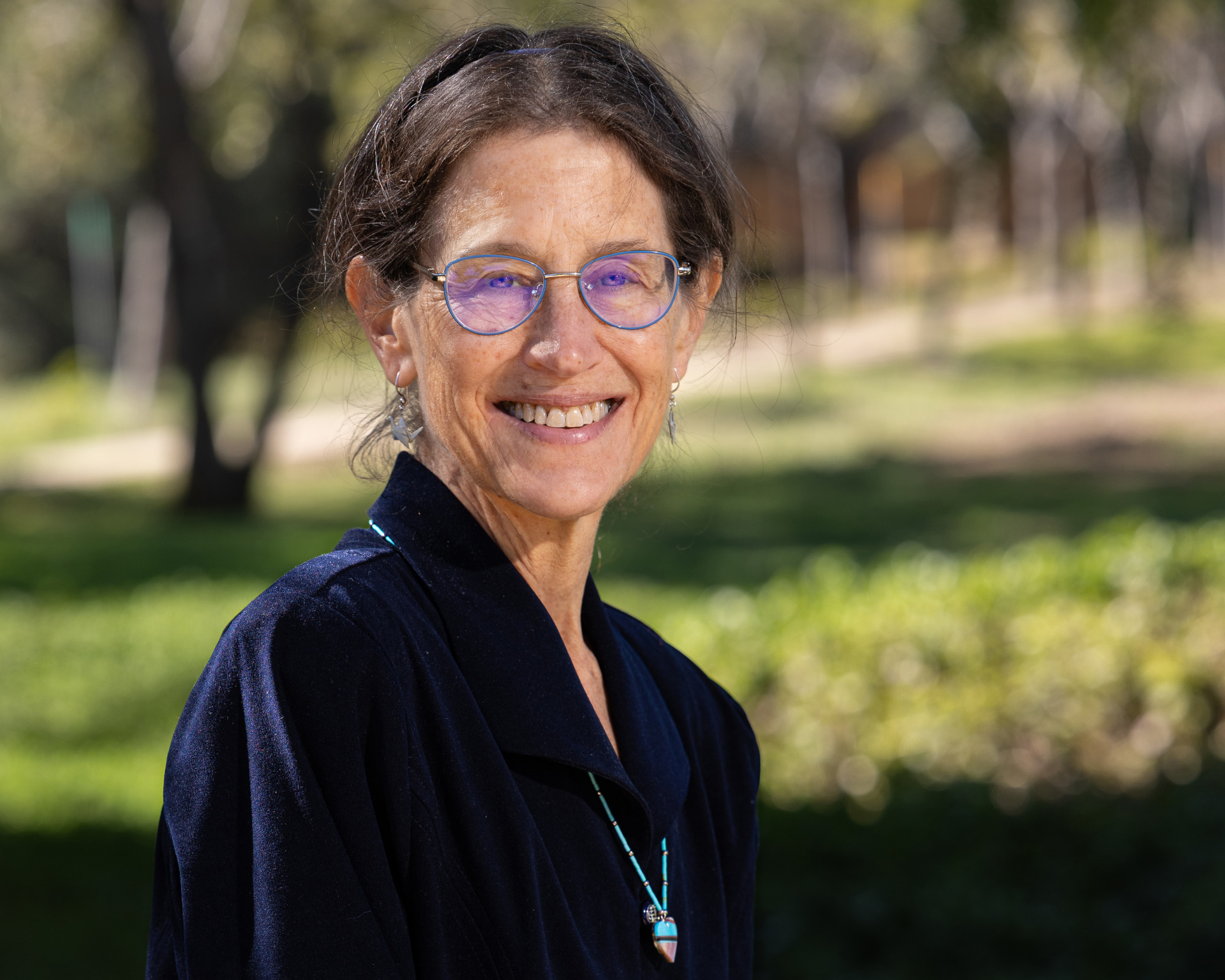A dark-haired woman wearing a navy blue collared shirt and blue-rimmed glasses smiles at the camera outside in a grassy area.