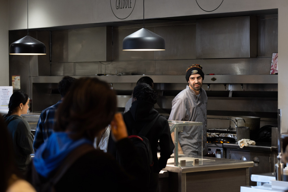 A man wearing a grey chef shirt turns away from a stove with foil items on it and smiles at students in line at the Bistro.