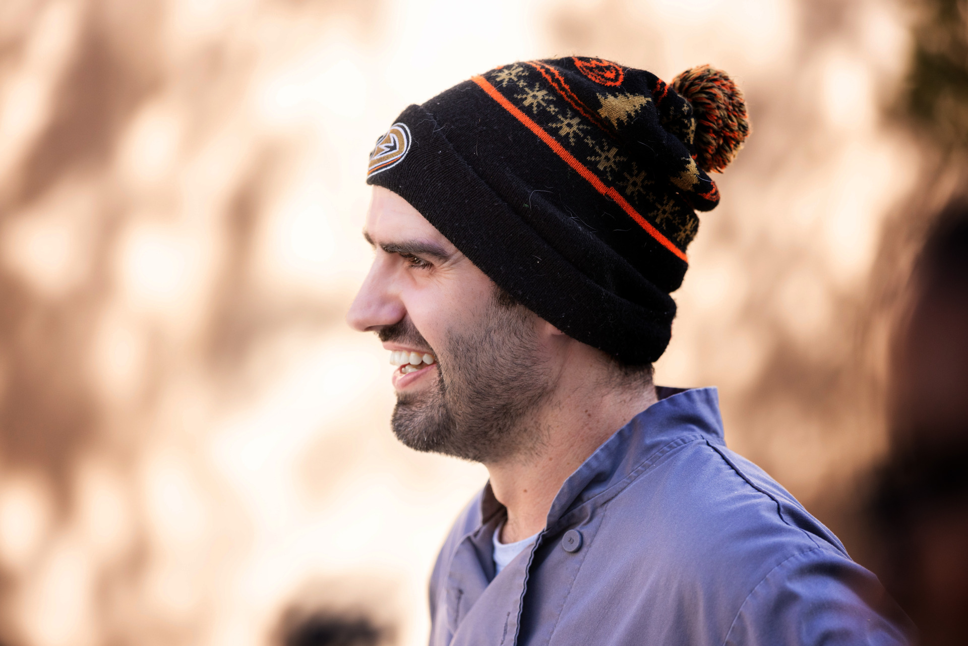 Side profile shot of a bearded man wearing a grey chef shirt and a black Anaheim Ducks holiday beanie smiles.