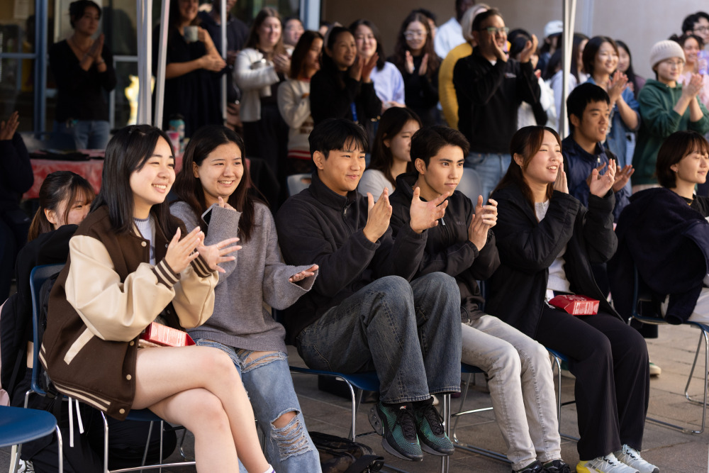 A large group of students, some sitting in chairs and standing in the background are clapping and smiling as they listen to someone out of frame.