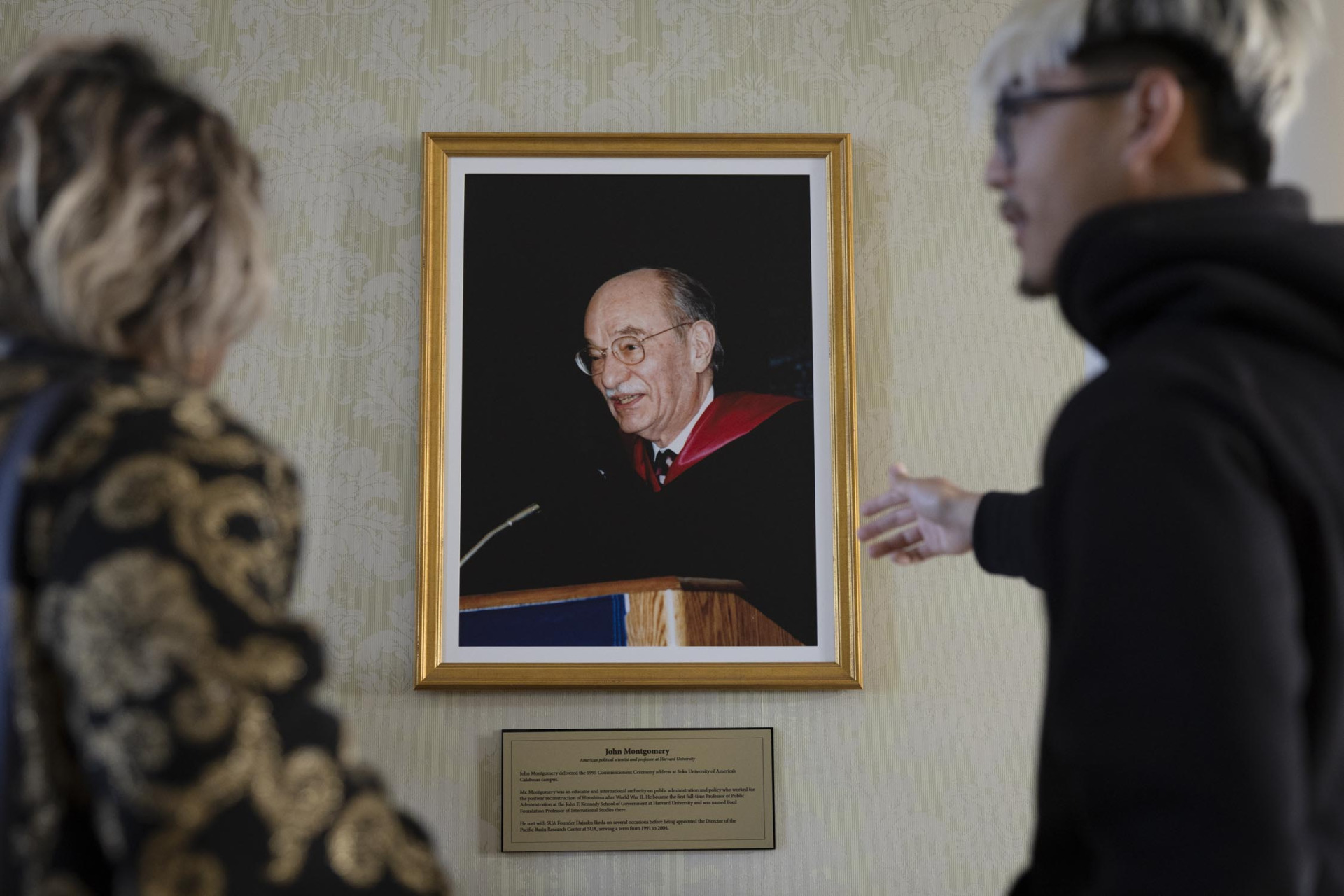 A male student with bleach blonde and black hair gestures towards a framed photo of a man speaking at a lectern.