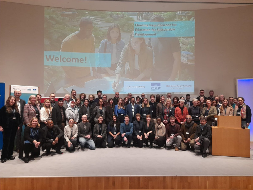 A large group of people in business casual attire stand in front of a welcome sign for a conference in Germany.