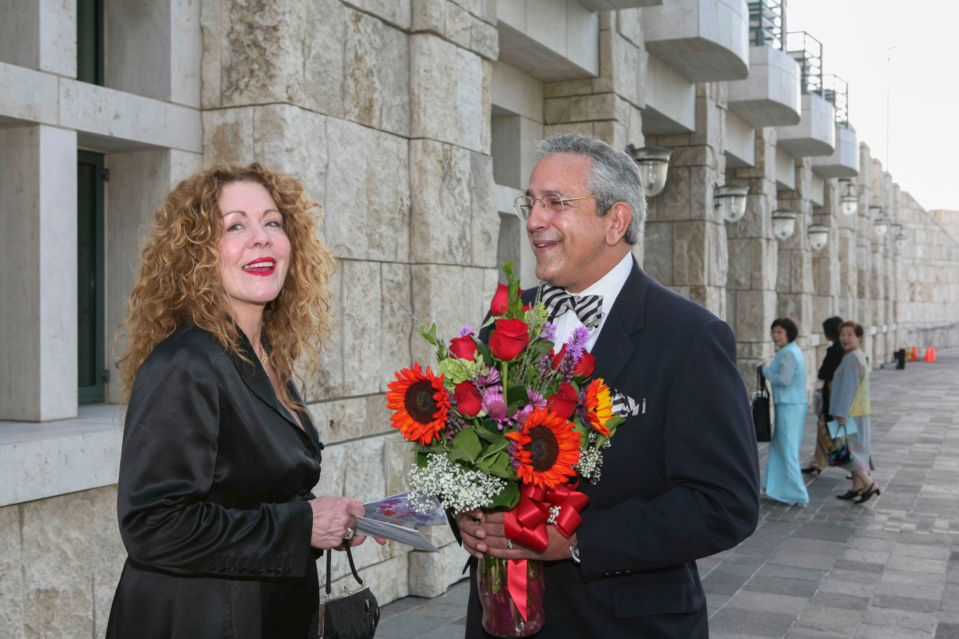 Linda and Luis Neieves are wearing black formal attire, smiling, and Luis is holding a bouquet of red and purple flowers as they stand outside of Founders Hall.