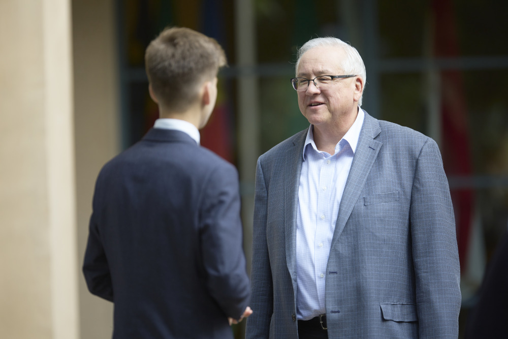 SUA President Ed Feasel is wearing a grey suit jacket and white shirt. He smiles as he speaks to a student wearing a blue suit. 