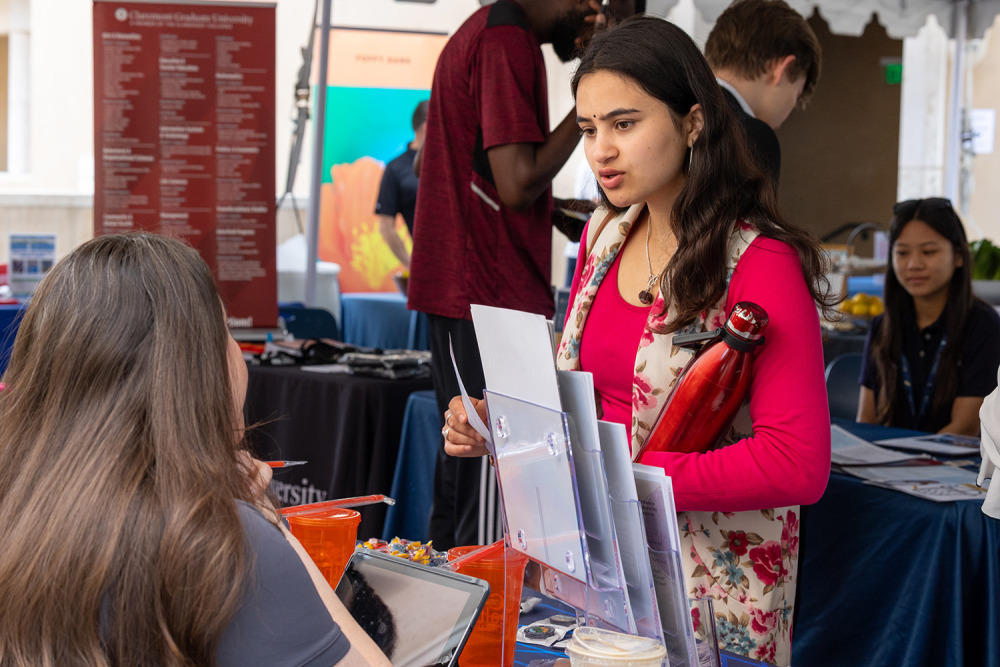 A student in a bright pink top and floral vest speaks with a recruiter at a career fair booth. She holds a red water bottle and a stack of papers, while a large informational banner for Claremont Graduate University stands behind them.