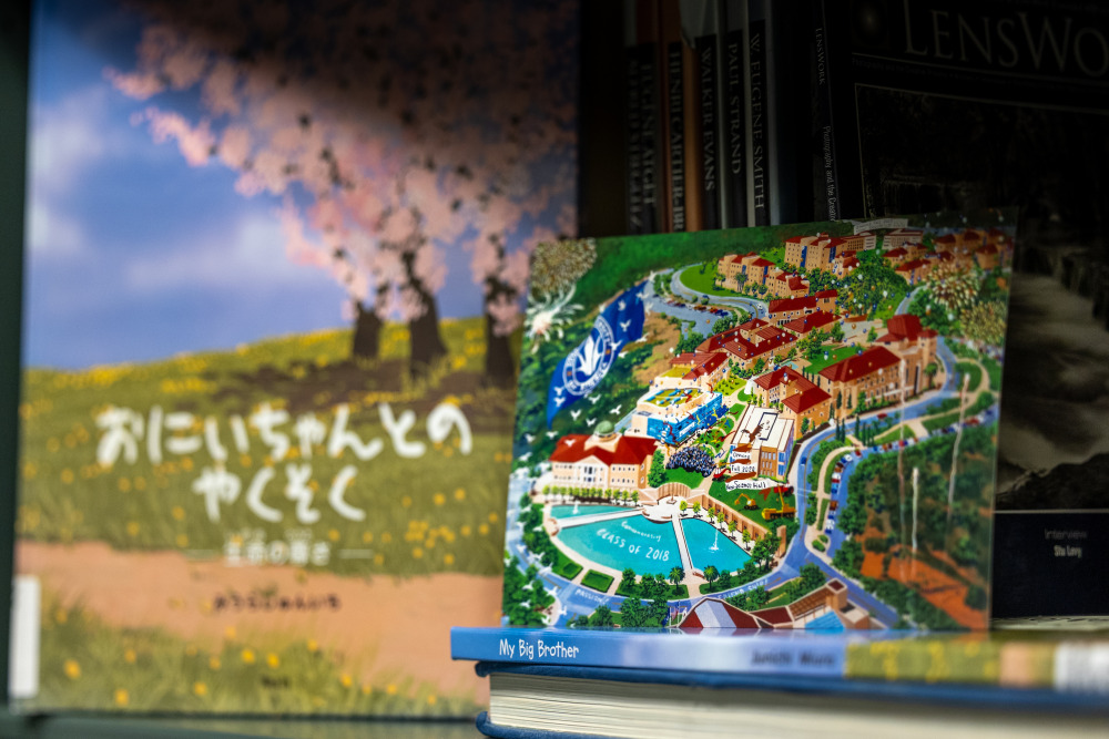 A children's book with cherry blossoms and Japanese characters on the cover is in the background of a postcard painting of an aerial view of SUA's campus.