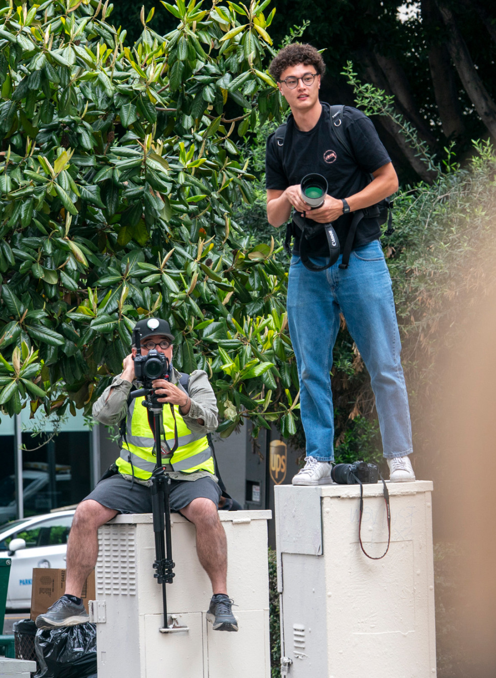 Two photographers perched on utility boxes outdoors. One man sits with a camera on a monopod, while a younger man with curly hair and glasses stands on the adjacent box, holding a camera with a large white telephoto lens.