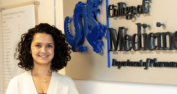 Aarohee Bhattarai has short curly dark brown hair and is wearing a white shirt. She is standing to the side of wall sign that says "College of Medicine"