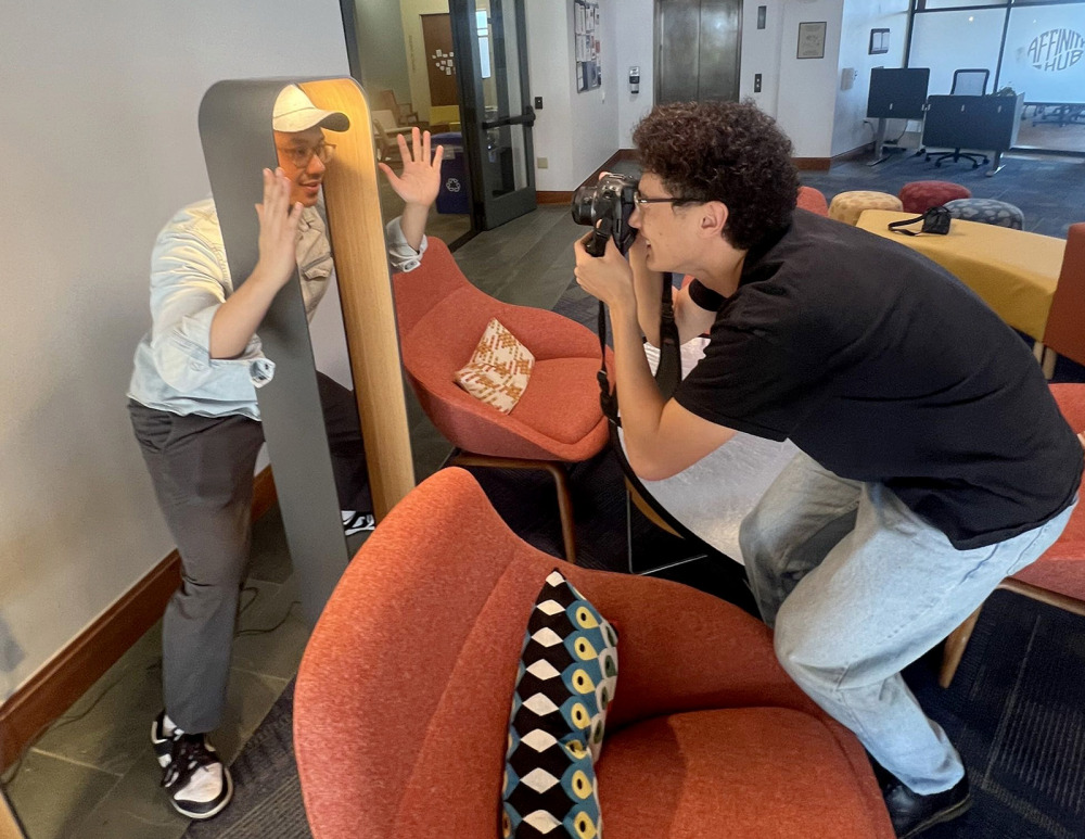 A photographer in a black T-shirt and light jeans leans in to take a photo of a man wearing a baseball cap who is playfully posing behind a modern, arched wooden floor lamp.