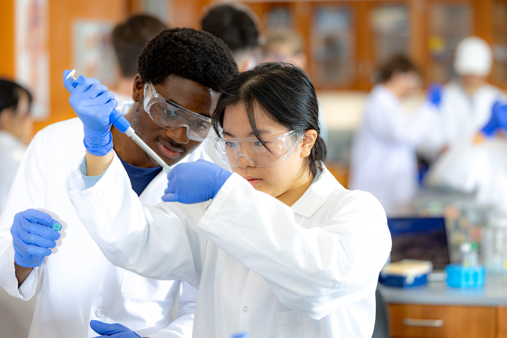 A male and female student in lab coats and safety goggles work together with a micropipette in a science lab.