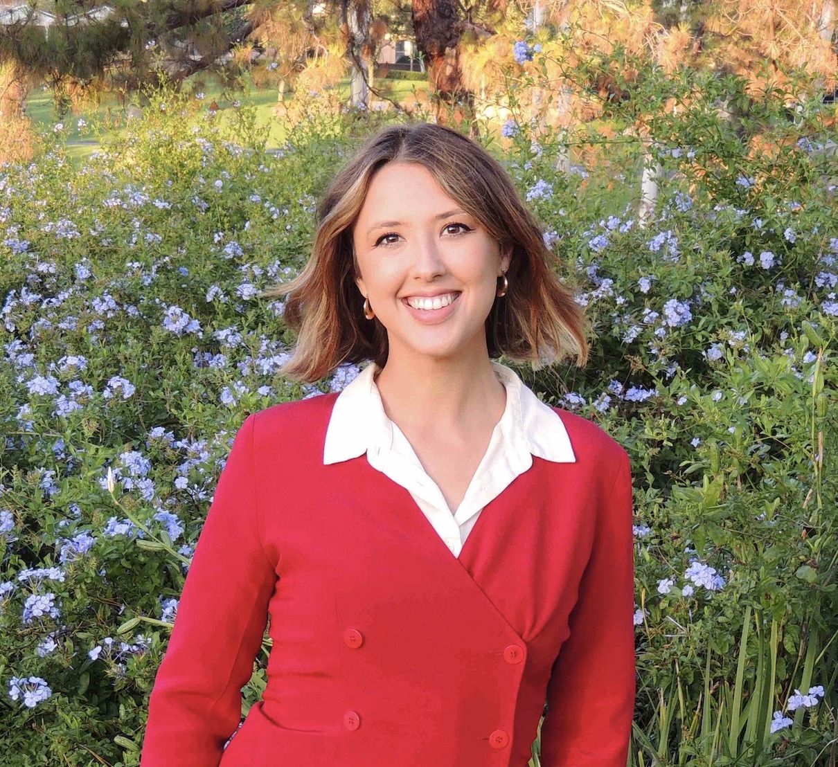 A woman with short brown hair wearing a red longsleeve shirt with a white collar poses in front of a field of blooming purple flowers.