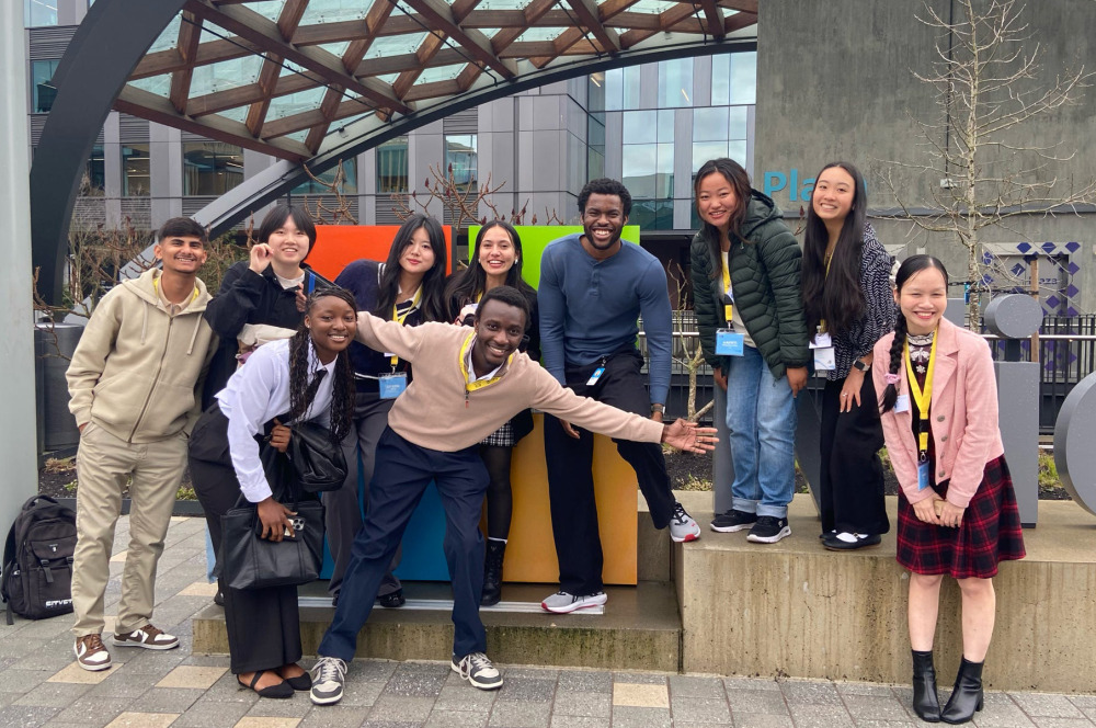A diverse group of young adults smile and pose together in front of a modern building with a glass and wood canopy. Several individuals are wearing identification lanyards.