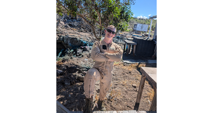 A man wearing a tan jumpsuit and sunglasses stands with his arms crossed and one boot up on a step as he works in an outdoor area.