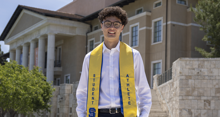 A man with curly brown hair is wearing a white dress shirt and yellow graduation stole that says "Student Athlete" as he stands in Front of a large building with white pillars.