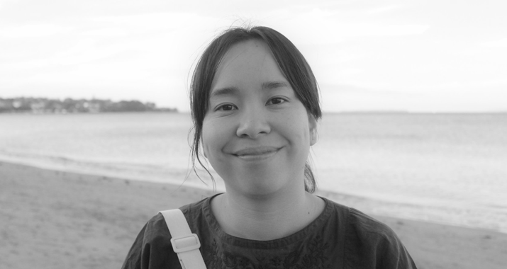 A woman with dark hair tied back in a ponytail stands in front of a body of water in a black and white photo.