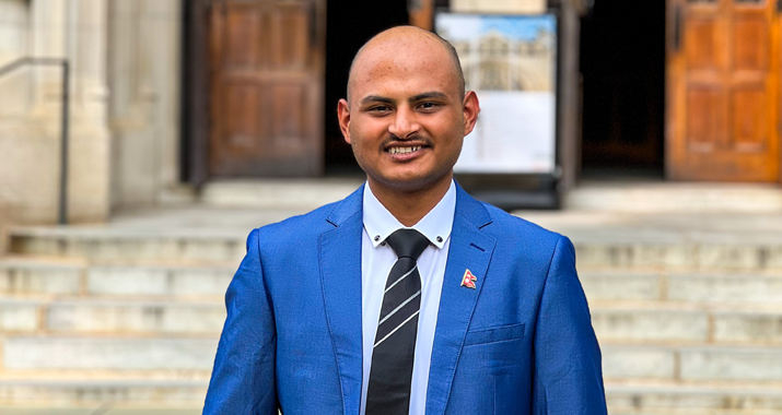 Mukesh Bastola wears a blue suit jacket and striped black tie as he stands in front of marble stairs outside a building with wooden doors.