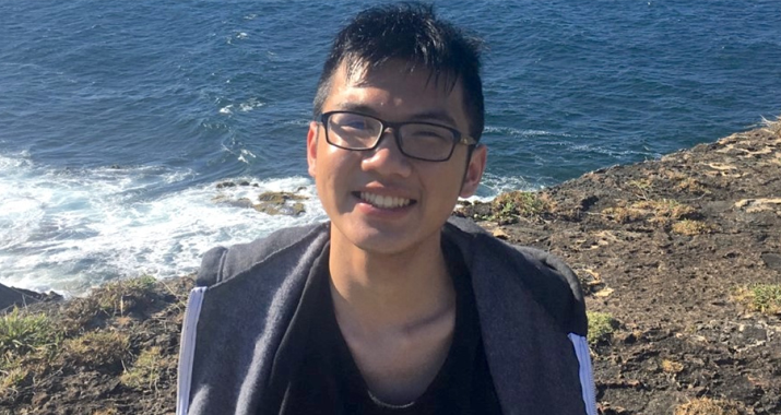 A man with short black hair and black eyeglasses smiles while standing on a cliffside overlooking the ocean