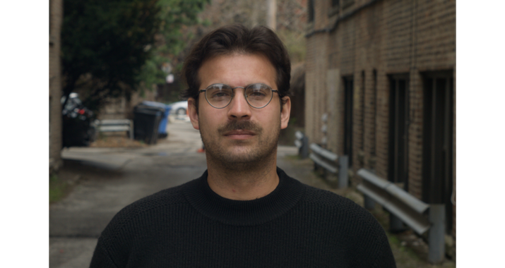 A man with short brown hair and facial hair wearing round glasses and a black longsleeve shirt poses in an alleyway lined with brick buildings.