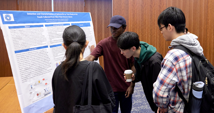 Three students look and listen to a fellow student presenting their summer research project on a posterboard.
