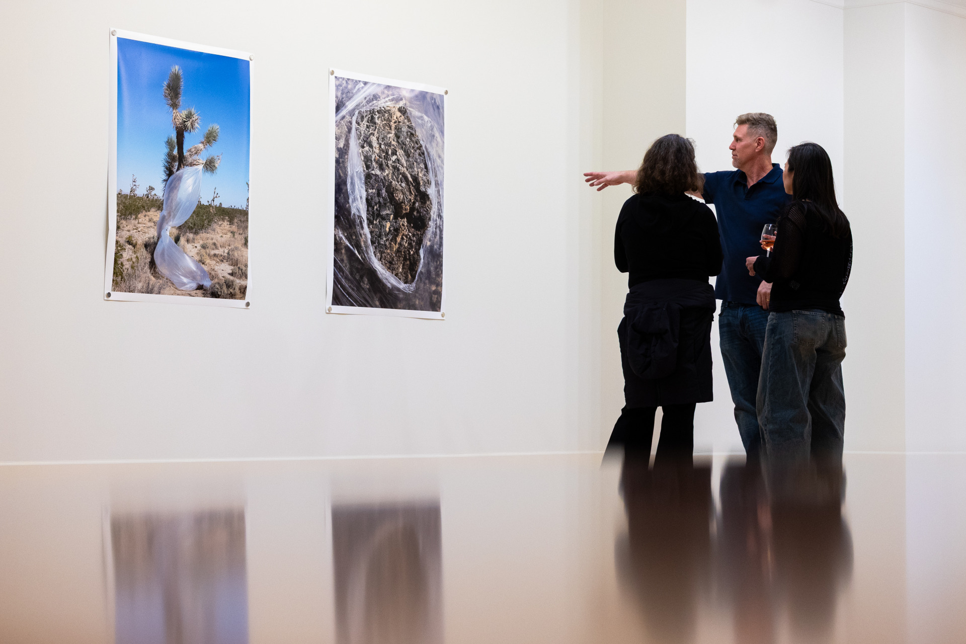 A man wearing a navy polo shirt is gesturing towards two large photographs on a white wall as he explains them to two women wearing black shirts.