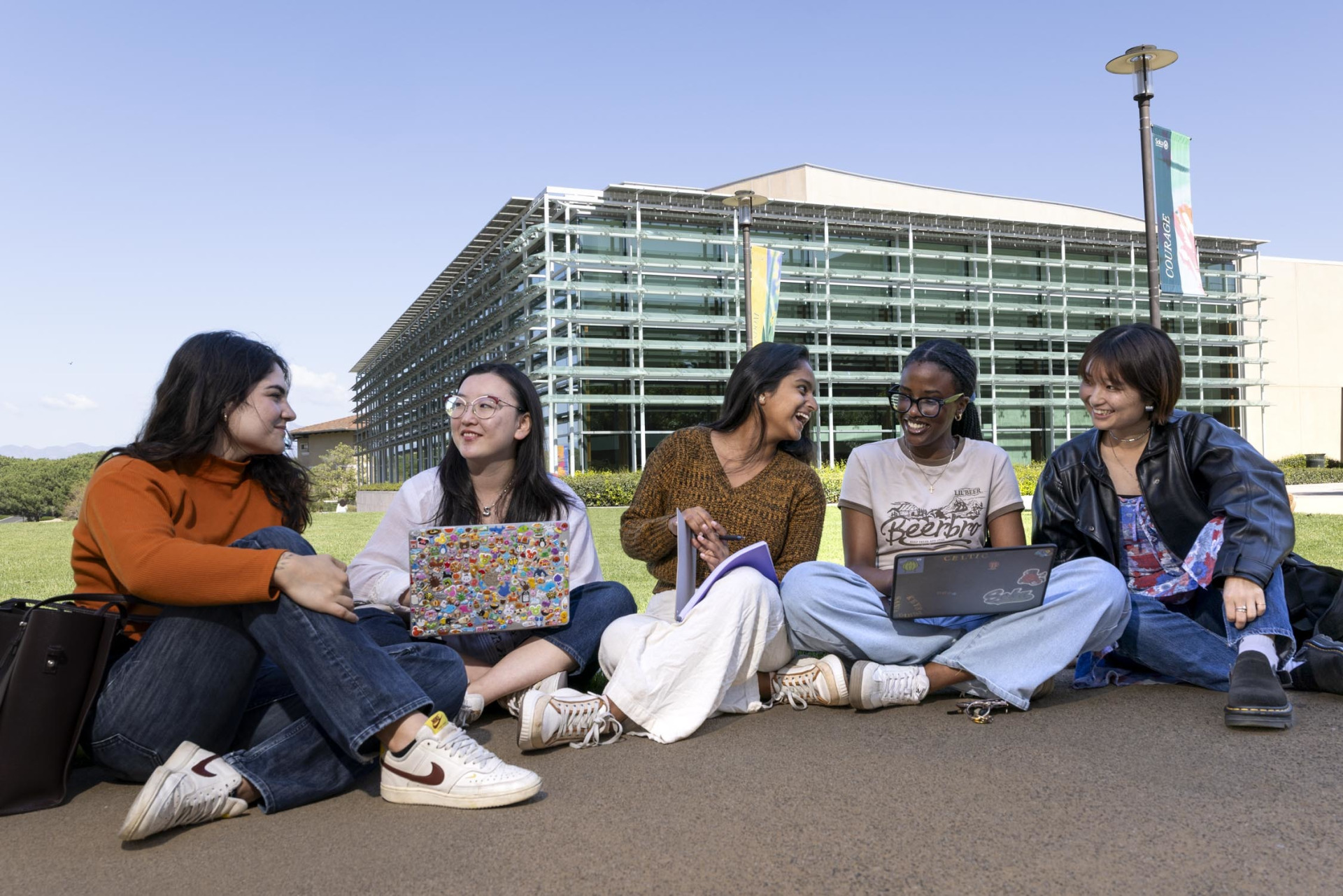 Five female students sit on the sidewalk in front of the Performing Arts Center, some with laptops in their laps, as they smile and laugh with eachother.