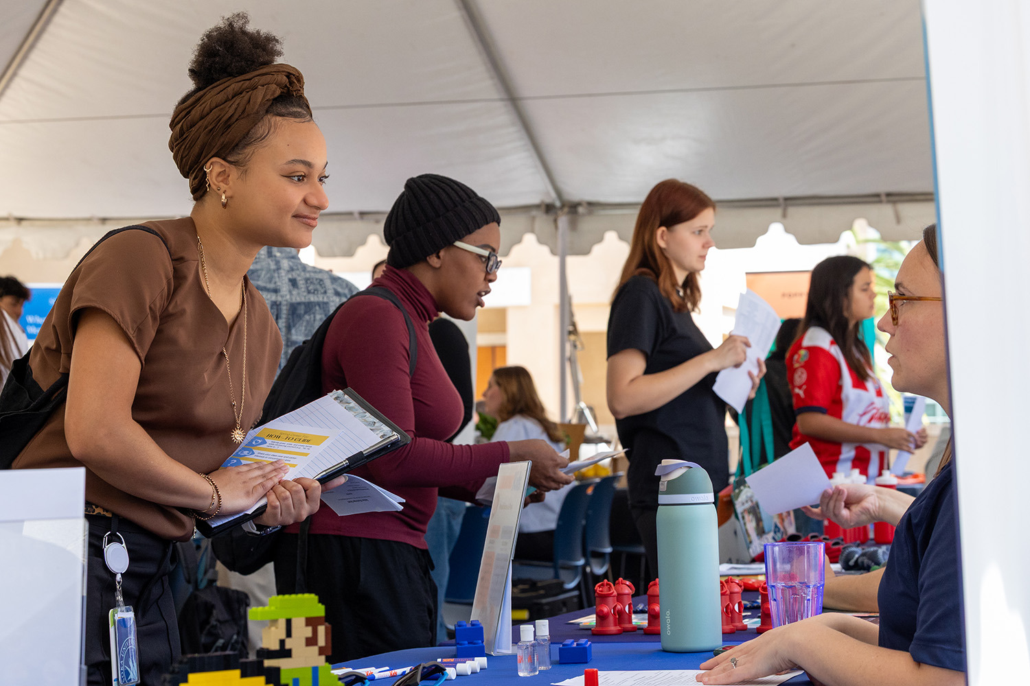 A young woman with a brown headwrap and gold jewelry smiles while leaning over a table at an outdoor career fair, holding a clipboard and documents. Other students and recruiters are visible under a large white tent in the background.