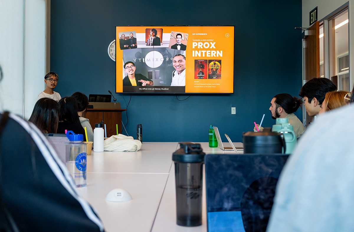 A student gives a presentation about an internship to a group seated at a long table. A large screen displays "PROX INTERN" and various photos.