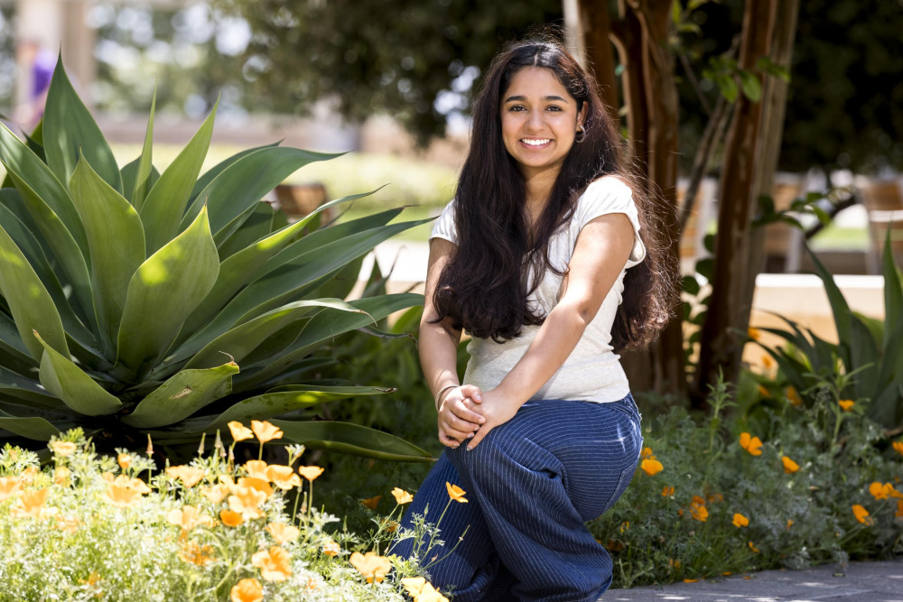 Aarna Marwa ’29 wears a white blouse and blue striped jeans as she kneels down by plants and flowers outdoor on SUA's campus.