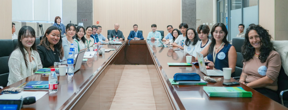 A group of students, staff, and faculty sit around the outside of a very long, rectangular table and look to the camera.