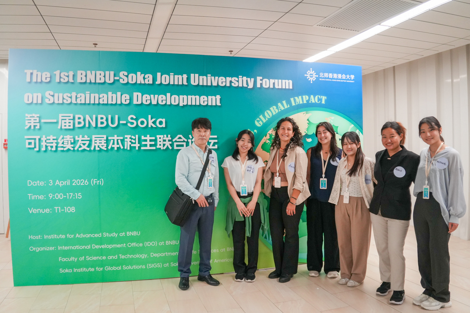 A man stands next to six women, all wearing nametags, as they pose in front of a large green and blue sign reading "The 1st BNBU-Soka Joint University Forum on Sustainable Development"