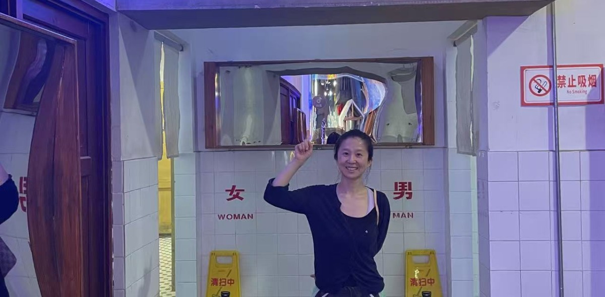 Dr. Yang Allen smiling and pointing upwards, standing in front of public restroom entrances with "WOMAN" and "MAN" signs.