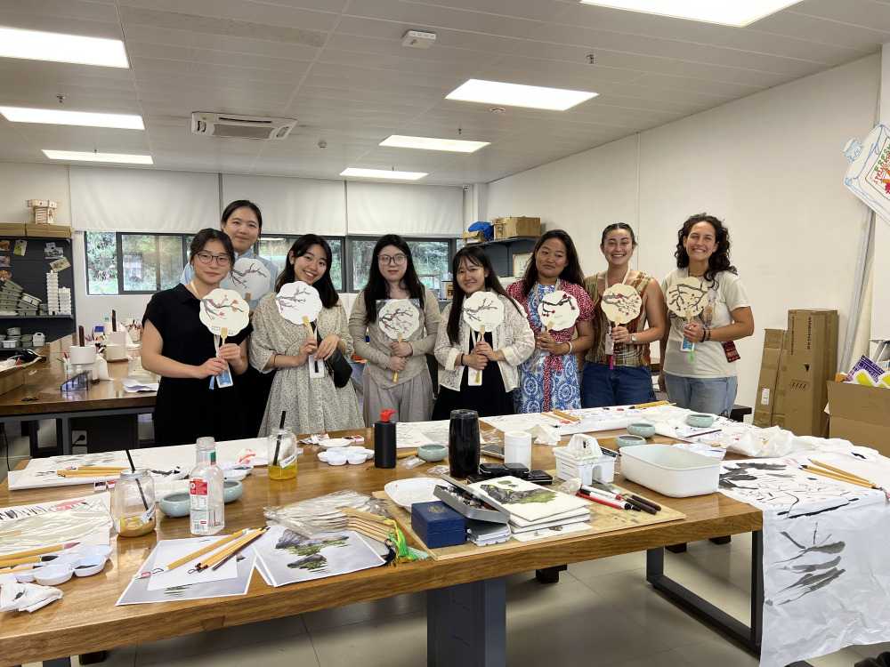 Eight women pose with similar artwork of cherry blossoms as they stand behind a wooden table full of art supplies including paint, papers, paintbrushes.